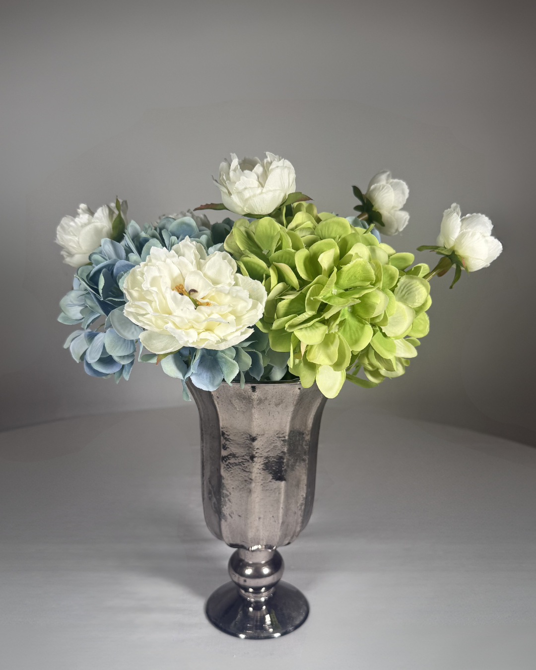 The Dakota silk hydrangea arrangement — three blue-and-green faux hydrangea stems with a single ivory peony in a clear glass vase. 11 inches tall. Hand-arranged in NYC by Deadass Flowers.