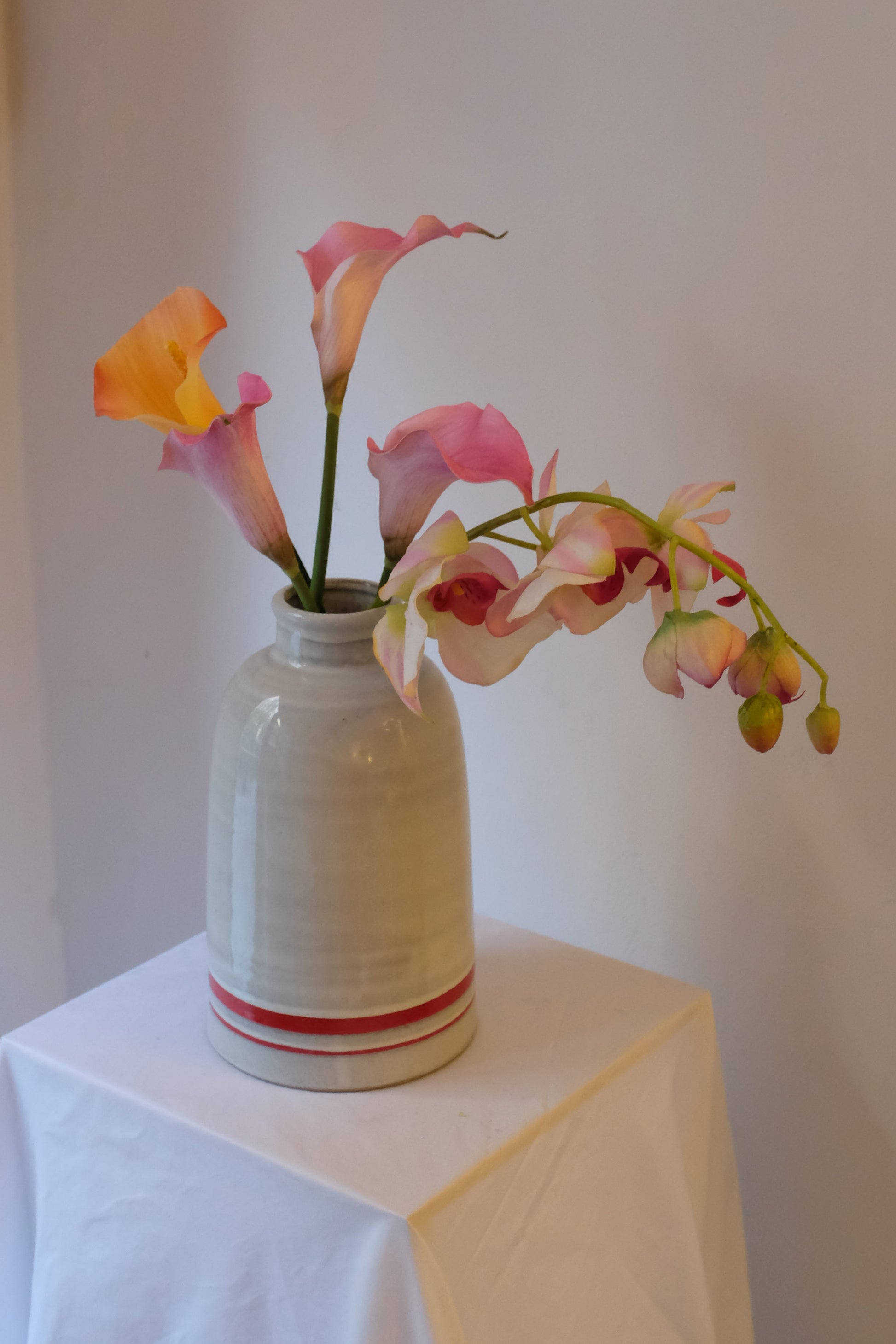 Callas + Orchid — orange and pink silk calla lilies with a trailing pink phalaenopsis orchid in a cream ceramic vintage bottle vessel.