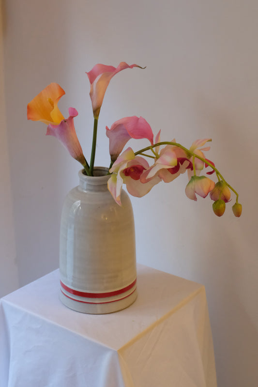 Callas + Orchid — orange and pink silk calla lilies with a trailing pink phalaenopsis orchid in a cream ceramic vintage bottle vessel.